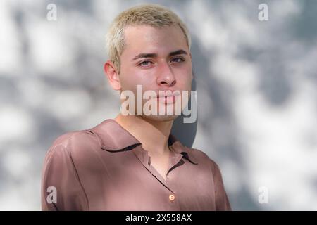 Porträt eines jungen Mannes mit blonden Haaren und einem braunen Hemd steht vor einer Wand. Er schaut direkt in die Kamera. Stockfoto