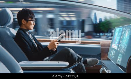 Futuristisches Konzept: Schöner, stilvoller japanischer Geschäftsmann in Brille, der Notizbuch liest und Nachrichten auf dem Augmented Reality Screen ansieht, während er in einem autonomen, selbstfahrenden emissionsfreien Auto sitzt. Stockfoto