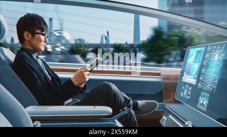 Futuristisches Konzept: Schöner, stilvoller japanischer Geschäftsmann in Brille, der Notizbuch liest und Nachrichten auf dem Augmented Reality Screen ansieht, während er in einem autonomen, selbstfahrenden emissionsfreien Auto sitzt. Stockfoto