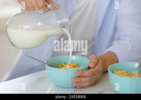 Frühstück machen. Mann, der Milch aus der Kanne in eine Schüssel mit Cornflakes auf weißem Marmortisch gießt, Nahaufnahme Stockfoto
