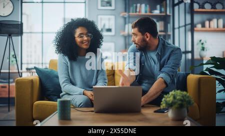 Verschiedene multiethnische Paare sitzen auf einer Couch Sofa in stilvollem Wohnzimmer und wählen Artikel aus, die online mit Laptop-Computer gekauft werden können. Freunde oder Kollegen diskutieren Arbeitsprojekte. Stockfoto