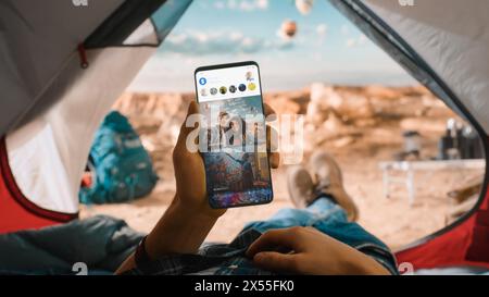 POV eines Touristen, der vertikal ein Smartphone hält und Social Network Feeds überprüft, Freunde neueste Fotos. Reisende, die sich in einem Zelt auf einem Rocky Mountain und fliegenden Heißluftballons ausruhen Stockfoto