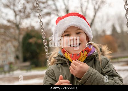 Freundliches, hübsches Mädchen auf einem Spielplatz mit einem schicken roten Weihnachtsmann-Hut. Im Winter, kurz vor Weihnachten, gibt es große Begeisterung über die Geschenke. Stockfoto