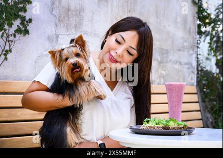 Fröhliche Dame mit einem Hund, der in einem Café im Freien sitzt, eine gesunde Mahlzeit und einen Smoothie genießt Stockfoto