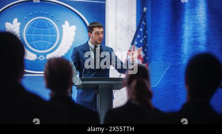 Junger Vertreter der Organisation beantwortet Fragen von Journalisten auf der Pressekonferenz. Pressesprecher, der beim Gipfeltreffen arbeitet. Minister im Gespräch mit dem Kongress. Hintergrund mit amerikanischen Flaggen. Stockfoto