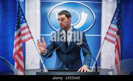 Junger Vertreter der Organisation beantwortet Fragen von Journalisten auf der Pressekonferenz. Pressesprecher, der beim Gipfeltreffen arbeitet. Minister im Gespräch mit dem Kongress. Hintergrund mit amerikanischen Flaggen. Stockfoto