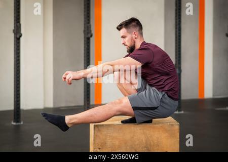 Seitenansicht eines starken bärtigen erwachsenen männlichen Athleten in Sportbekleidung, der wegschaut, während er auf der Box sitzt und Calisthenics-Pistolenhockübung bei Tageslicht macht Stockfoto