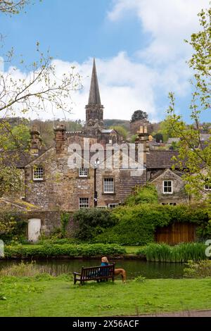 Frau, die auf einer Bank am Ufer des Flusses Wye sitzt, in der Stadt Bakewell im Peak District Derbyshire, mit Hütten und der Kirche der Allerheiligen Stockfoto