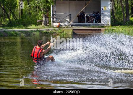 Ein Wakeboarder stürzt auf einem Wasserbrett entlang des Ufers eines Waldsees durch das Wasser und erzeugt wunderschöne Wasserspritzer in seiner Nähe. Stockfoto