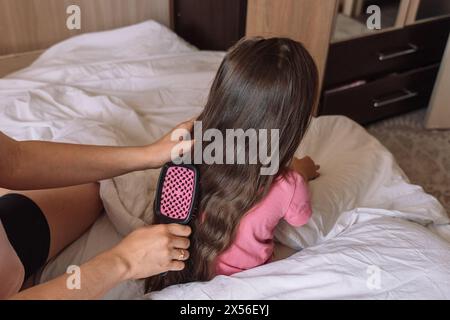 Mom kämmt das Haar ihrer Tochter mit einem Kamm, während sie im Bett sitzt Stockfoto