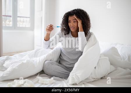 Junge Frau, die ihre Temperatur überprüft, während sie im Bett sitzt Stockfoto