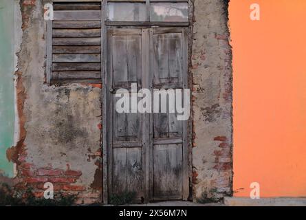 251 völlig verblasste Holztür auf einer abgebrochenen, nicht verputzten Mauerwand zwischen anderen grünen und orangen Fassaden, im Plaza Mayor Square. Trinidad-Kuba. Stockfoto