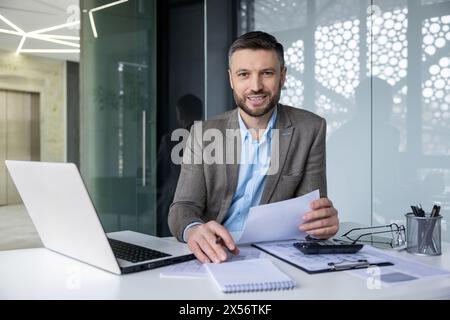Selbstbewusster Geschäftsmann mittleren Alters in einem stilvollen, modernen Büro, das an seinem Schreibtisch arbeitet. Er liest Dokumente, während er mit einem Laptop interagiert, und zeigt Professionalität und Konzentration. Stockfoto