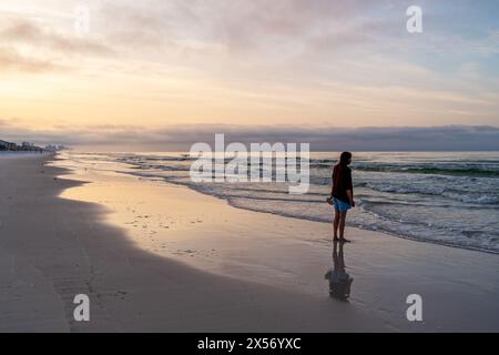Barfüßige Frau, die Schuhe hält und bei Sonnenaufgang auf das Meer, das Meer oder den Golf von Mexiko blickt. Stockfoto