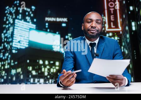 afroamerikanischer Gastgeber, der Nachrichten während der späten Abendshow präsentiert und internationale Schlagzeilen des Tages liest. Man Journalist, der sich mit aktuellen Nachrichten über die neuesten Ereignisse befasst. Stockfoto