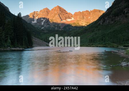 Aufsteigendes Sonnenlicht auf den Maroon Bells in der Mitte des Sommers mit Maroon Lake im Vordergrund. Die Maroon Bells befinden sich in der Nähe der Stadt Aspen Colorado. Stockfoto