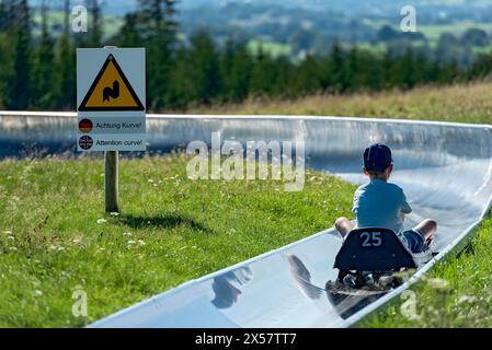 Junge mit Rodeln auf der Sommerrodelbahn, Warnschild Aufmerksamkeitskurve, Ski- und Rodelarena, Hoherodskopf Berggipfel, Ausflug Stockfoto