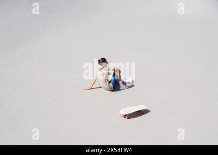 Ein junger Mann macht eine Pause vom Skateboarden und sitzt beiläufig auf dem Boden mit seinem Surfskate in der Nähe. Das helle Sonnenlicht wirft einen scharfen Schatten auf den Th Stockfoto