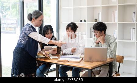 Eine reife asiatische Lehrerin hält ihren Schülern im Klassenzimmer einen Vortrag, erklärt die letzten Projektdetails, hilft ihren Schülern, Stockfoto