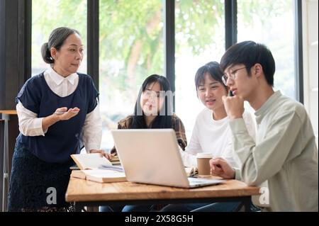Eine reife asiatische Lehrerin hält ihren Schülern im Klassenzimmer einen Vortrag, erklärt die letzten Projektdetails, hilft ihren Schülern, Stockfoto