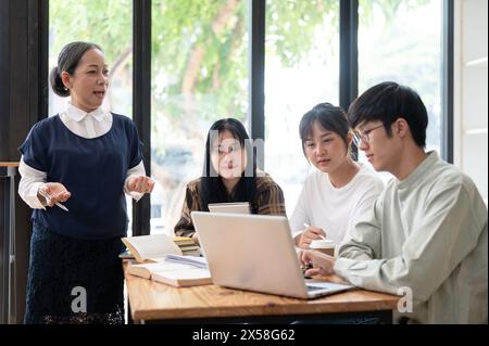 Eine reife asiatische Lehrerin hält ihren Schülern im Klassenzimmer einen Vortrag, erklärt die letzten Projektdetails, hilft ihren Schülern, Stockfoto
