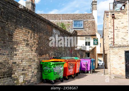 Farbenfrohe Abfalleimer, die an einer Ziegelmauer entlang Miltons Walk zur Cut Throat Alley mit Graffiti angereiht sind. Cambridge, England, Großbritannien Stockfoto