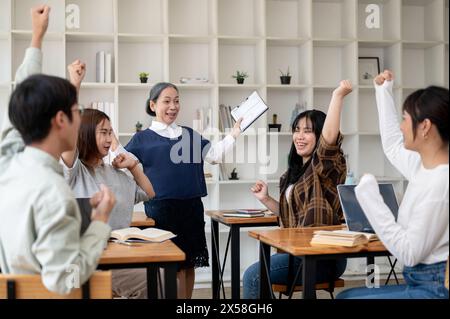 Eine fröhliche, reife asiatische Lehrerin oder Professorin, die gute Nachrichten mit Schülern im Klassenzimmer feiert. Bildungskonzept Stockfoto