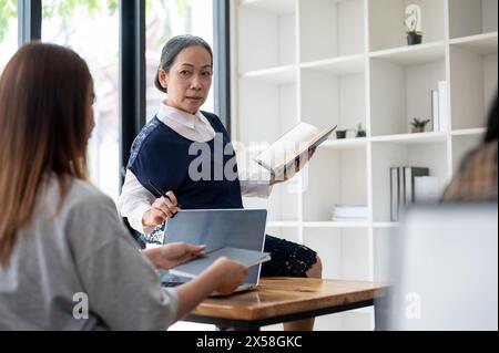 Eine professionelle, reife asiatische Lehrerin oder Professorin sitzt auf einem Tisch, während sie ihren Schülern im Klassenzimmer einen Vortrag hält und eine Lektion gibt. Ed Stockfoto