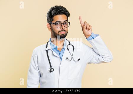 Eureka. Inspirierter junger indischer Arzt Kardiologe Mann, der mit offenem Mund den Finger nach oben zeigt, gute Idee, Plan, Start-up, Inspiration Motivation Lösung hat. Apotheke-Typ auf beigefarbenem Hintergrund Stockfoto