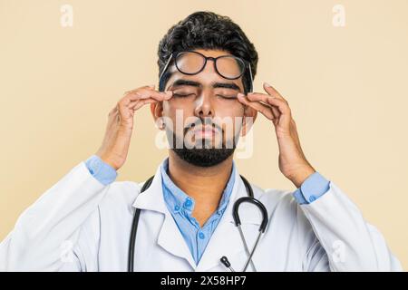 Erschöpft müde indische junge Doktor Kardiologe Mann zieht die Brille aus, fühlt Augenschmerzen, ist Überarbeit Burnout von langer Arbeit. Schläfriger, erschöpfter Kerl, der die Augen isoliert auf beigefarbenem Hintergrund reibt Stockfoto