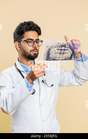 Der indische junge Arzt Kieferorthopäden untersucht ein Panorama-Röntgenbild der Kieferzähne. 3D-Modell des Mundes des Patienten, MRT-Scan. Zahnheilkunde, Mundpflege. Ein arabischer Stomatologe auf beigefarbenem Hintergrund Stockfoto