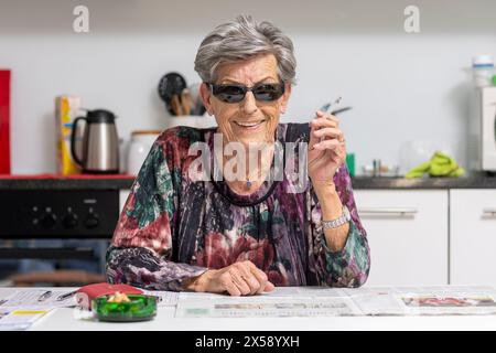 Eine alte Frau mit Falten sitzt in der Küche mit Sonnenbrille und raucht fröhlich eine Zigarette. Stockfoto