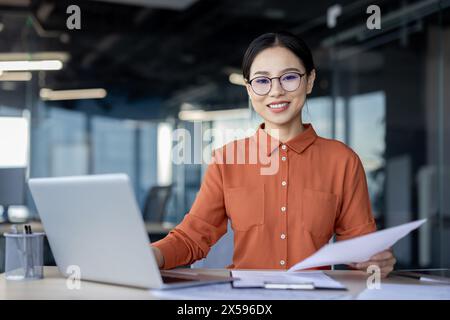 Eine selbstbewusste Geschäftsfrau mit Brille lächelt, während sie Dokumente hält. Sie arbeitet in einem dynamischen Büroumfeld mit offenem Laptop neben ihr. Stockfoto