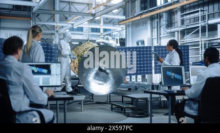 Techniker in Schutzanzügen, die an Satellitenkonstruktionen arbeiten, unter der Kontrolle des Chefingenieurs. Fertigungseinrichtung Der Luftfahrtbehörde: Wissenschaftler, Die Raumfahrzeuge Montieren. Mission Zur Weltraumforschung Stockfoto