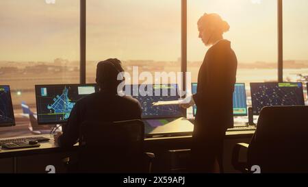 Vielseitiges Air Traffic Control Team arbeitet im modernen Airport Tower bei Sonnenuntergang. Die Controller feiern den ersten Start eines neuen Verkehrsflugzeugs in einem Raum voller Computer mit Handklatschen. Stockfoto