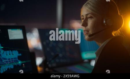 Nahaufnahme weibliches Porträt der Fluglotse mit Headset bei einem Anruf im Flughafenturm bei Nacht sprechen. Der Büroraum ist voll von Computerdisplays mit Navigationsbildschirmen, Flugradardaten. Stockfoto