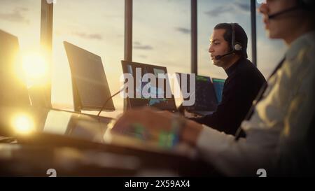 Männlicher Fluglotse mit Headset sprechen bei einem Anruf im Airport Tower. Der Büroraum ist voll mit Desktop-Computerdisplays mit Navigationsbildschirmen, Flugzeugabflug- und Ankunftsdaten für das Team. Stockfoto