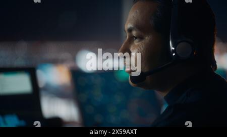 Nahaufnahme Porträt eines männlichen Fluglotsen mit Headset-Gespräch bei einem Anruf im Airport Tower. Der Büroraum ist voll von Desktop-Computer-Displays mit Navigationsbildschirmen, Flugradardaten. Stockfoto