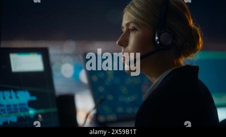 Nahaufnahme weibliches Porträt der Fluglotse mit Headset bei einem Anruf im Flughafenturm bei Nacht sprechen. Der Büroraum ist voll von Computerdisplays mit Navigationsbildschirmen, Flugradardaten. Stockfoto