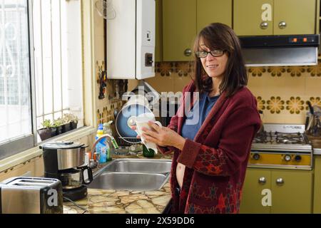 Eine Frau steht in einer Küche vor dem Fenster und trocknet einen gewaschenen Becher. Die Küche ist überfüllt mit verschiedenen Geräten und Utensilien, Stockfoto