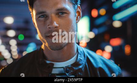 Porträt eines gutaussehenden ernsthaften Mannes, der in die Kamera schaut, stehend in der Nachtstadt mit Bokeh Neon Street Lights im Hintergrund. Selbstbewusster junger Mann mit stilvollem Haar. Nahaufnahme Stockfoto