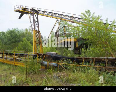 Der verdrehte und rostfreie Kadaver des Förderbandes in einem verlassenen Steinbruch, wo die Vegetation alles bedeckt hat. Stockfoto