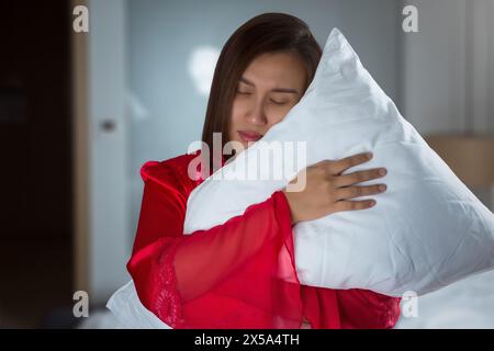 Eine asiatische Frau in einem roten Seidennachthemd schläft beim Stehen ein und umarmt nachts ein Kissen in ihrem Schlafzimmer. Stockfoto