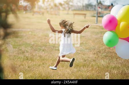 Glücklich, Mädchen und Running im Feld mit Ballons von Karneval, Markt und Festival für Erfolg, Feiern oder Spielen. Aktiv, Kind und Sprung im Sommer Stockfoto