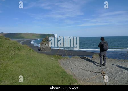 Junger erwachsener Mann, der die großartige Aussicht auf den schwarzen Sandstrand von Reynisfjara und die Umgebung von der Spitze von Dyrhólaey, Süd-Island, bewundert Stockfoto