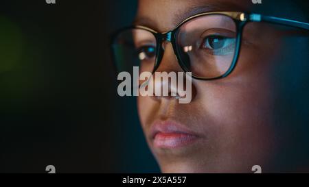 Nahaufnahme Porträt eines niedlichen jungen Teenagers multiethnische Schwarze Mädchen in einem dunklen gemütlichen Zimmer zu Hause. Die junge Frau trägt eine Brille, die die Hausaufgaben von ihrem Laptop-Bildschirm reflektiert. Stockfoto