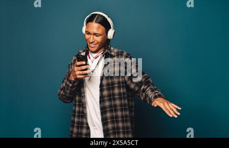 Glücklicher, lässiger Mann mit geflochtenen Haaren steht in einem Studio. Er hört Musik auf seinem Handy mit blauem Hintergrund. Stockfoto