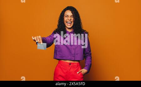 Die fröhliche junge Frau steht auf einem orangefarbenen Hintergrund und hält eine Bankkarte. Sie tippt, um mit einem fröhlichen Lächeln zu bezahlen, und bezahlt mühelos bargeldlos. Stockfoto