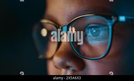 Nahaufnahme Porträt eines niedlichen jungen Teenagers multiethnische Schwarze Mädchen in einem dunklen gemütlichen Zimmer zu Hause. Die junge Frau trägt eine Brille, die die Hausaufgaben von ihrem Laptop-Bildschirm reflektiert. Stockfoto