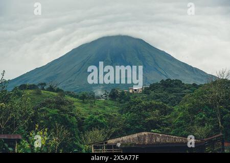 Majestätischer Blick auf den Vulkan Arenal, umgeben von Wolken mit grüner Landschaft und einem Haus in Costa Rica Stockfoto